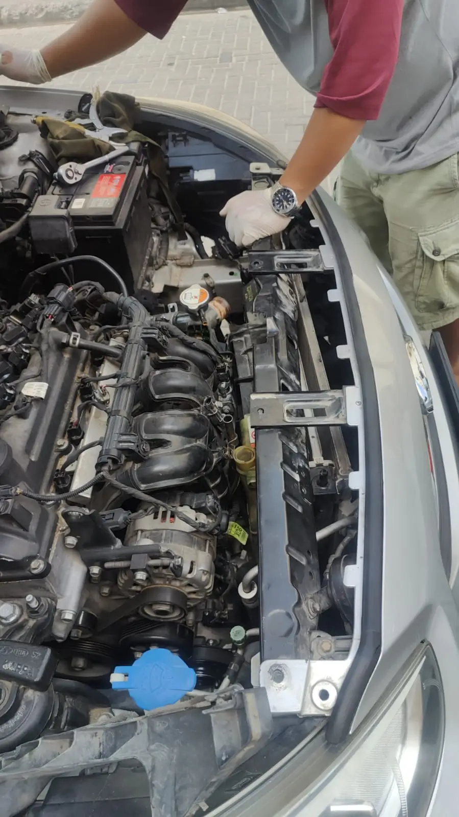 Mechanic with gloves working on open car engine bay, inspecting radiator and alternator